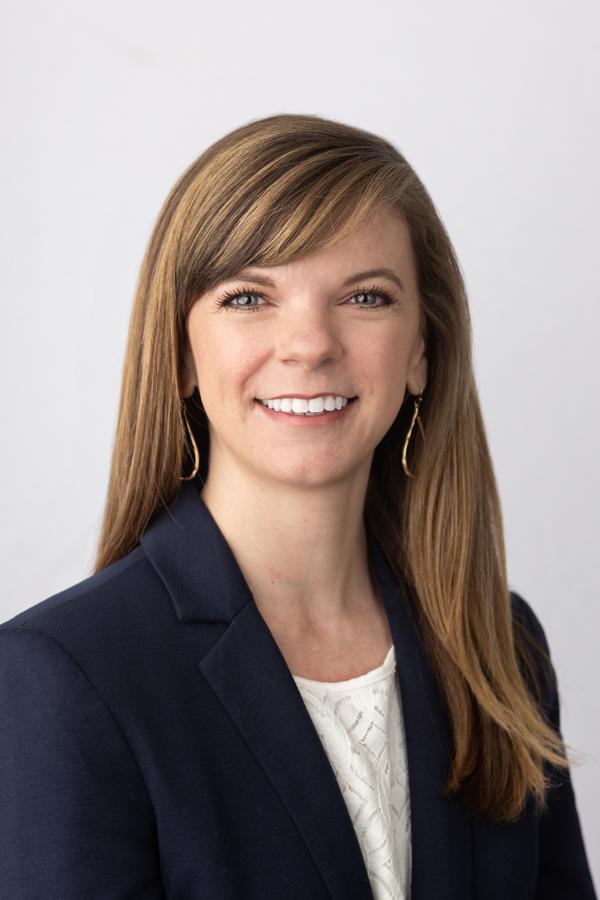 Image of Dr. Courtney East, who has long brown hair with highlights and a smile, wearing a navy suit jacket over a lace shirt. The background is white. 